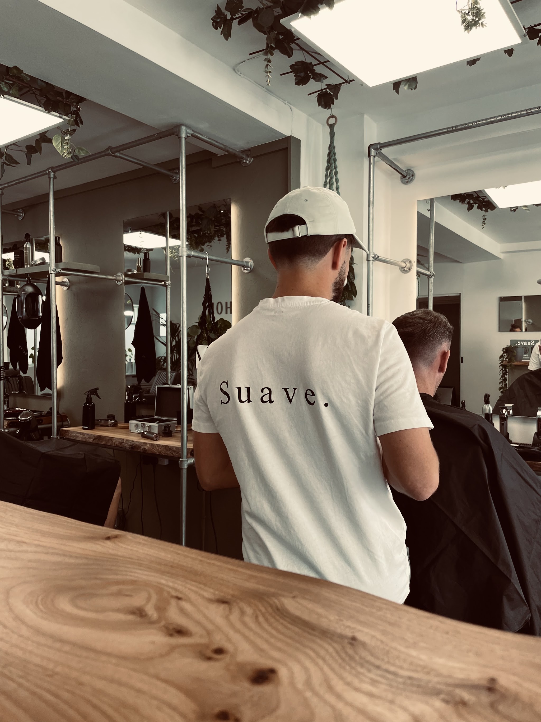 Barber in a white Suave. T-shirt cutting a client's hair beside the live-edge wood counter